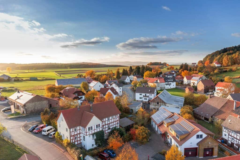 Aerial view of a picturesque rural village with traditional houses, surrounded by lush green fields and colorful autumn trees, showcasing scenic landscape and peaceful community living.